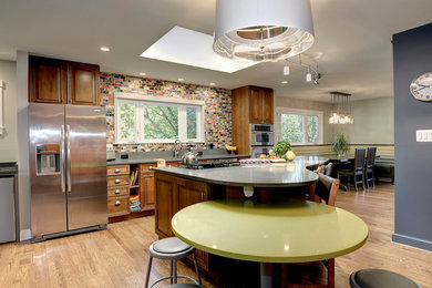 Mid-sized eclectic single-wall light wood floor and brown floor eat-in kitchen photo in DC Metro with raised-panel cabinets, medium tone wood cabinets, solid surface countertops, multicolored backsplash, ceramic backsplash, stainless steel appliances, an island and an undermount sink
