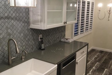 Photo of a mid-sized transitional galley separate kitchen in Los Angeles with a farmhouse sink, glass-front cabinets, white cabinets, granite benchtops, grey splashback, ceramic splashback, stainless steel appliances, light hardwood floors, no island, grey floor and grey benchtop.