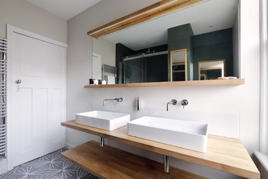 This is an example of a mid-sized contemporary kids bathroom in Sussex with open cabinets, medium wood cabinets, a curbless shower, a wall-mount toilet, green tile, stone tile, beige walls, ceramic floors, an integrated sink and wood benchtops.