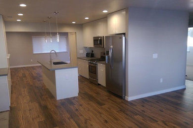 Example of a mid-sized minimalist galley medium tone wood floor and brown floor open concept kitchen design in Other with a drop-in sink, flat-panel cabinets, white cabinets, quartz countertops, stainless steel appliances, an island and gray countertops