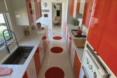 На фото: отдельная, параллельная кухня среднего размера в стиле ретро с одинарной мойкой, плоскими фасадами, оранжевыми фасадами, белым фартуком, фартуком из керамической плитки, белой техникой, разноцветным полом и белой столешницей без острова с
