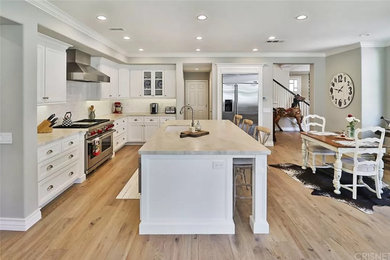 Inspiration for a large country l-shaped open plan kitchen in Los Angeles with an undermount sink, shaker cabinets, white cabinets, quartzite benchtops, white splashback, porcelain splashback, stainless steel appliances, medium hardwood floors, with island, brown floor and beige benchtop.
