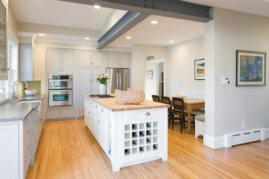 Mid-sized trendy l-shaped light wood floor eat-in kitchen photo in San Francisco with a farmhouse sink, recessed-panel cabinets, gray cabinets, wood countertops, white backsplash, ceramic backsplash, stainless steel appliances and an island