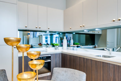 This is an example of a mid-sized contemporary l-shaped open plan kitchen in London with a drop-in sink, flat-panel cabinets, beige cabinets, marble benchtops, metallic splashback, mirror splashback, stainless steel appliances, light hardwood floors and no island.