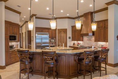 Example of a huge tuscan u-shaped travertine floor open concept kitchen design in Miami with a farmhouse sink, raised-panel cabinets, brown cabinets, granite countertops, beige backsplash, stone tile backsplash, stainless steel appliances and two islands