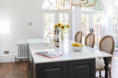 This is an example of a mid-sized traditional l-shaped open plan kitchen in London with a farmhouse sink, shaker cabinets, black cabinets, quartzite benchtops, white splashback, ceramic splashback, black appliances, dark hardwood floors, with island, brown floor and white benchtop.
