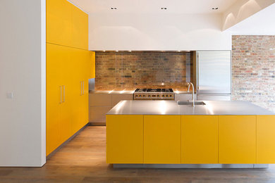 Design ideas for a large urban galley kitchen/diner in London with an integrated sink, flat-panel cabinets, yellow cabinets, stainless steel worktops, multi-coloured splashback, glass tiled splashback, stainless steel appliances, laminate floors, an island and brown floors.