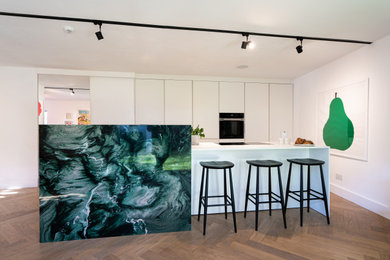 Photo of a medium sized contemporary galley open plan kitchen in London with a built-in sink, flat-panel cabinets, white cabinets, quartz worktops, green splashback, engineered quartz splashback, black appliances, light hardwood flooring, an island, brown floors, white worktops and a drop ceiling.