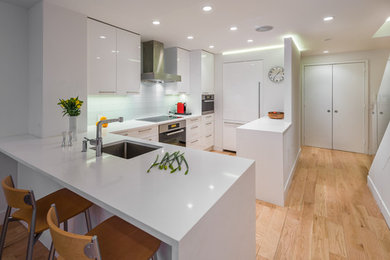 Design ideas for a mid-sized modern galley eat-in kitchen in New York with a drop-in sink, flat-panel cabinets, white cabinets, solid surface benchtops, white splashback, glass tile splashback, white appliances, light hardwood floors and a peninsula.