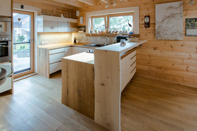 Design ideas for a mid-sized country l-shaped open plan kitchen in Munich with flat-panel cabinets, beige cabinets, wood benchtops, beige splashback, light hardwood floors, a drop-in sink, stainless steel appliances, a peninsula, brown floor, brown benchtop and glass sheet splashback.