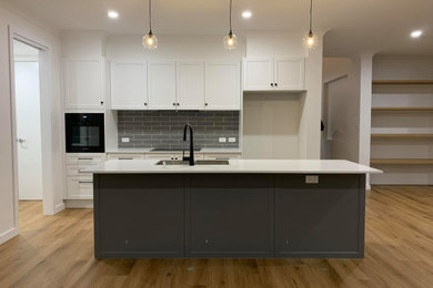 Inspiration for a mid-sized galley open plan kitchen in Adelaide with a double-bowl sink, shaker cabinets, solid surface benchtops, grey splashback, subway tile splashback, black appliances, vinyl floors, with island, brown floor and white benchtop.