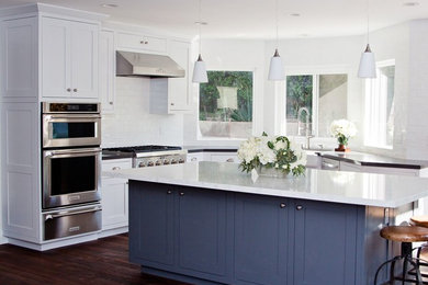 Example of a mid-sized transitional l-shaped dark wood floor and brown floor kitchen design in Los Angeles with a farmhouse sink, shaker cabinets, white cabinets, white backsplash, subway tile backsplash, stainless steel appliances and an island