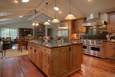 Example of a large country u-shaped medium tone wood floor and brown floor open concept kitchen design in Other with a farmhouse sink, raised-panel cabinets, medium tone wood cabinets, granite countertops, black backsplash, stone slab backsplash, stainless steel appliances and an island