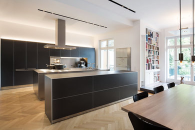 Offene, Geräumige Moderne grifflose Küche mit Doppelwaschbecken, flächenbündigen Schrankfronten, schwarzen Schränken, Edelstahl-Arbeitsplatte, Küchenrückwand in Metallic, Elektrogeräten mit Frontblende, braunem Holzboden und zwei Kücheninseln in Köln