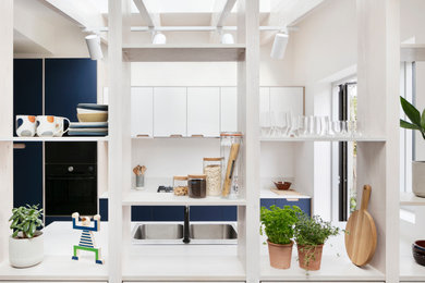 This is an example of a medium sized scandinavian grey and white galley open plan kitchen in London with a built-in sink, flat-panel cabinets, blue cabinets, composite countertops, white splashback, granite splashback, black appliances, porcelain flooring, no island, grey floors, white worktops, exposed beams and feature lighting.