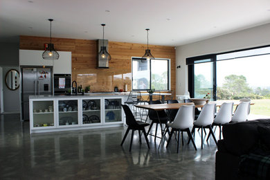 This is an example of a mid-sized modern single-wall eat-in kitchen in Auckland with a farmhouse sink, flat-panel cabinets, white cabinets, solid surface benchtops, brown splashback, glass sheet splashback, stainless steel appliances, concrete floors, with island and grey floor.
