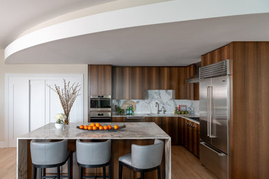 Cette image montre une cuisine ouverte en L avec des portes de placard marrons, une crédence blanche, un électroménager en acier inoxydable, parquet clair, îlot, un sol marron et un plan de travail blanc.