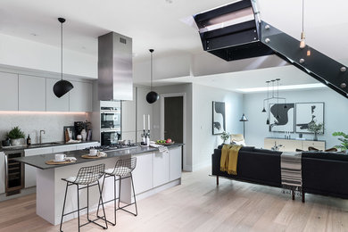 Design ideas for a contemporary galley open plan kitchen in London with a submerged sink, flat-panel cabinets, white cabinets, grey splashback, light hardwood flooring, an island, beige floors and black worktops.