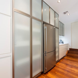 Réalisation d'une arrière-cuisine minimaliste de taille moyenne avec un placard à porte vitrée, une façade en inox, un plan de travail en quartz modifié, une crédence blanche, une crédence en feuille de verre, un sol en bois clair, un sol marron et un plan de travail blanc.