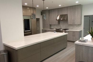 Inspiration for a mid-sized transitional l-shaped separate kitchen in Los Angeles with flat-panel cabinets, grey cabinets, marble benchtops, white splashback, stone slab splashback, stainless steel appliances, light hardwood floors and multiple islands.