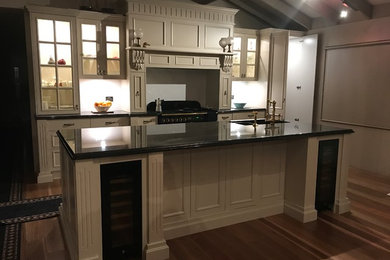 Photo of a mid-sized traditional galley kitchen pantry in Wollongong with an undermount sink, recessed-panel cabinets, white cabinets, granite benchtops, white splashback, ceramic splashback, black appliances, medium hardwood floors, with island, brown floor and multi-coloured benchtop.