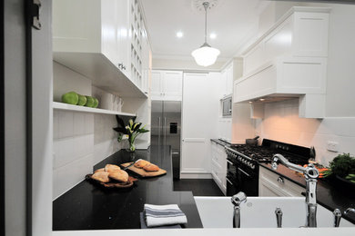 Photo of a traditional u-shaped separate kitchen with a farmhouse sink, shaker cabinets, white cabinets, solid surface benchtops, white splashback, ceramic splashback, black appliances, painted wood floors, no island, black floor and black benchtop.