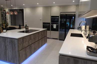 Design ideas for a medium sized contemporary galley kitchen/diner in Kent with a submerged sink, flat-panel cabinets, medium wood cabinets, quartz worktops, black splashback, glass tiled splashback, black appliances, ceramic flooring, an island, white floors and white worktops.