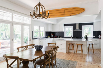 Photo of a large beach style u-shaped eat-in kitchen in Perth with a drop-in sink, shaker cabinets, white cabinets, solid surface benchtops, black splashback, metal splashback, stainless steel appliances, medium hardwood floors, with island and white benchtop.