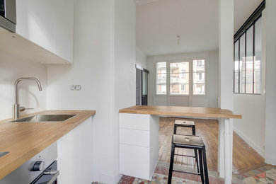 Cette photo montre une petite cuisine encastrable moderne en U fermée avec un évier 1 bac, un placard à porte plane, des portes de placard blanches, un plan de travail en bois, une crédence blanche, un sol multicolore et un plan de travail marron.