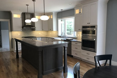 Inspiration for a transitional l-shaped light wood floor kitchen remodel in Boston with a farmhouse sink, shaker cabinets, white cabinets, granite countertops, gray backsplash, stone tile backsplash, stainless steel appliances and an island