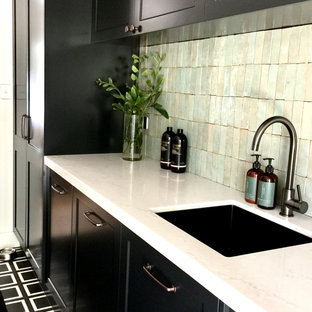 Aménagement d'une buanderie linéaire campagne dédiée et de taille moyenne avec un évier encastré, un placard à porte shaker, des portes de placard noires, un plan de travail en quartz modifié, une crédence verte, une crédence en carreau de terre cuite, un mur blanc, béton au sol, des machines côte à côte, un sol noir et un plan de travail blanc.