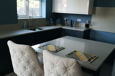 This is an example of a medium sized kitchen/diner in Kent with a built-in sink, flat-panel cabinets, blue cabinets, quartz worktops, blue splashback, black appliances, laminate floors, an island, grey floors and white worktops.