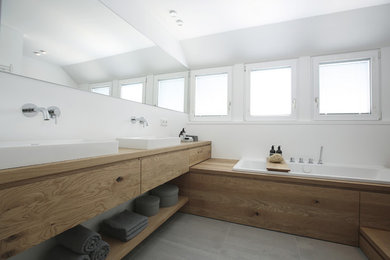Mid-sized contemporary bathroom in Frankfurt with flat-panel cabinets, light wood cabinets, a drop-in tub, gray tile, white walls, a vessel sink, wood benchtops and grey floor.