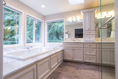 Inspiration for a mid-sized traditional master bathroom in Seattle with beaded inset cabinets, beige cabinets, a corner tub, a corner shower, white tile, stone slab, grey walls, light hardwood floors, an undermount sink, quartzite benchtops, grey floor, a hinged shower door and white benchtops.