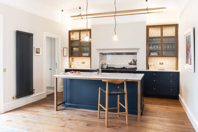 This is an example of a medium sized traditional single-wall kitchen/diner in Edinburgh with a submerged sink, shaker cabinets, blue cabinets, engineered stone countertops, white splashback, ceramic splashback, black appliances, light hardwood flooring, an island and white worktops.