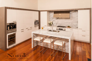Mid-sized trendy l-shaped medium tone wood floor eat-in kitchen photo in New Orleans with an undermount sink, flat-panel cabinets, white cabinets, marble countertops, white backsplash, stone slab backsplash, stainless steel appliances and an island