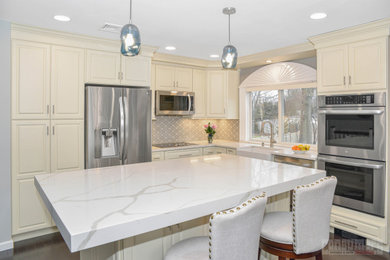 This is an example of a mid-sized transitional l-shaped eat-in kitchen in New York with a farmhouse sink, raised-panel cabinets, yellow cabinets, quartz benchtops, grey splashback, stainless steel appliances, with island and white benchtop.
