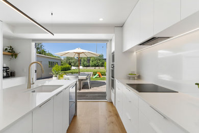Design ideas for a large contemporary single-wall open plan kitchen in Auckland with an undermount sink, flat-panel cabinets, white cabinets, quartz benchtops, white splashback, engineered quartz splashback, stainless steel appliances, medium hardwood floors, with island, brown floor and white benchtop.