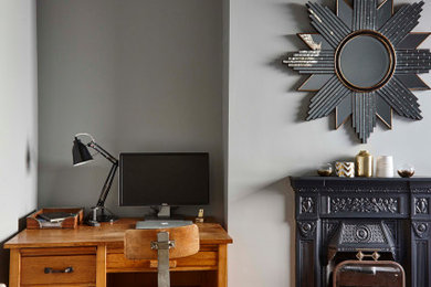 Modelo de despacho ecléctico pequeño con paredes grises, suelo de madera clara, todas las chimeneas, marco de chimenea de metal, escritorio independiente y suelo beige