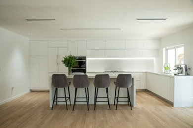 Large minimalist l-shaped medium tone wood floor and brown floor open concept kitchen photo in Miami with an undermount sink, flat-panel cabinets, white cabinets, quartz countertops, white backsplash, quartz backsplash, paneled appliances, an island and white countertops