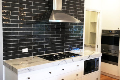 This is an example of a large modern galley open plan kitchen in Sydney with a double-bowl sink, flat-panel cabinets, white cabinets, marble benchtops, black splashback, subway tile splashback, with island and white benchtop.