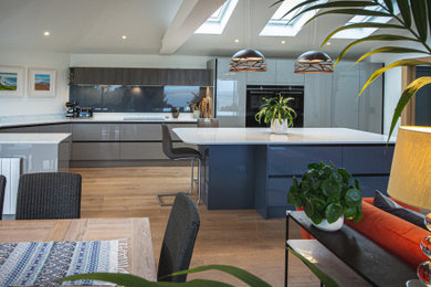 Photo of a large nautical grey and white l-shaped open plan kitchen in Devon with an integrated sink, flat-panel cabinets, blue cabinets, quartz worktops, grey splashback, glass sheet splashback, black appliances, light hardwood flooring, an island, brown floors, white worktops, a vaulted ceiling and feature lighting.