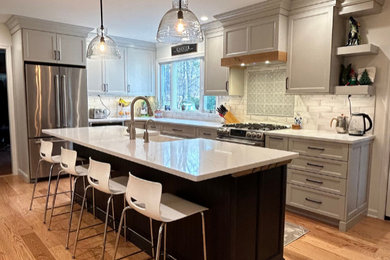 Large transitional l-shaped light wood floor and blue floor eat-in kitchen photo in New York with a farmhouse sink, recessed-panel cabinets, gray cabinets, quartz countertops, gray backsplash, ceramic backsplash, stainless steel appliances, an island and white countertops