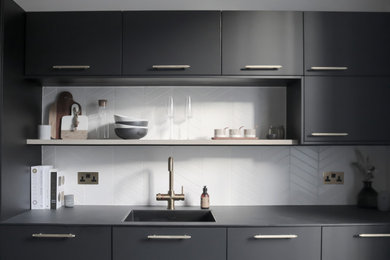 This is an example of a medium sized scandinavian single-wall enclosed kitchen in Edinburgh with a submerged sink, flat-panel cabinets, black cabinets, composite countertops, white splashback, ceramic splashback, black appliances, light hardwood flooring, an island, beige floors, black worktops and feature lighting.