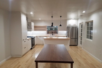 This is an example of a mid-sized contemporary l-shaped eat-in kitchen in Los Angeles with a farmhouse sink, flat-panel cabinets, white cabinets, wood benchtops, white splashback, stainless steel appliances, light hardwood floors, with island and ceramic splashback.