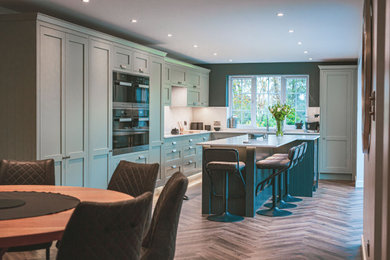 Photo of a medium sized traditional l-shaped open plan kitchen in Devon with an integrated sink, shaker cabinets, green cabinets, quartz worktops, black appliances, medium hardwood flooring, an island, brown floors, white worktops and feature lighting.