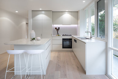 Photo of a mid-sized contemporary kitchen in Melbourne with a drop-in sink, flat-panel cabinets, beige cabinets, granite benchtops, white splashback, stone slab splashback, stainless steel appliances, light hardwood floors, with island and beige benchtop.
