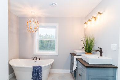 Mid-sized double-sink, ceramic tile and beige floor freestanding bathtub photo in Other with raised-panel cabinets, blue cabinets, a one-piece toilet, wood countertops, black countertops, a built-in vanity, gray walls and a vessel sink
