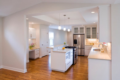 Inspiration for a mid-sized transitional l-shaped medium tone wood floor eat-in kitchen remodel in Austin with a double-bowl sink, flat-panel cabinets, white cabinets, solid surface countertops, beige backsplash, stone tile backsplash, stainless steel appliances and an island