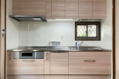 他の地域にあるおしゃれなI型キッチン (中間色木目調キャビネット、ステンレスカウンター、白いキッチンパネル、シルバーの調理設備) の写真